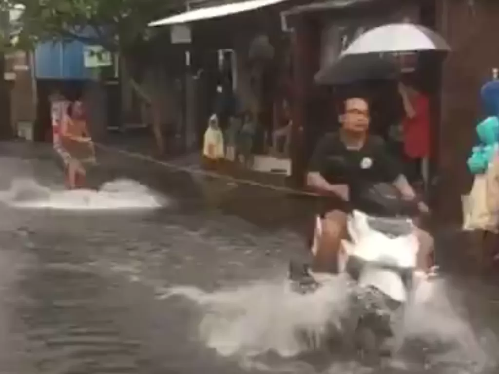 Tampilan pria yang tengah ditarik oleh pemotor dan melakukan atraksi surfing di jalanan banjir. (SS/Twitter/@Mom_isback)