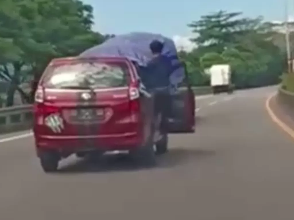 Tampilan pria yang berkendara mobil sambil bergelantungan di pintu mobil ketika berada di jalan tol. (SS/Instagram/@makassar_iinfo)