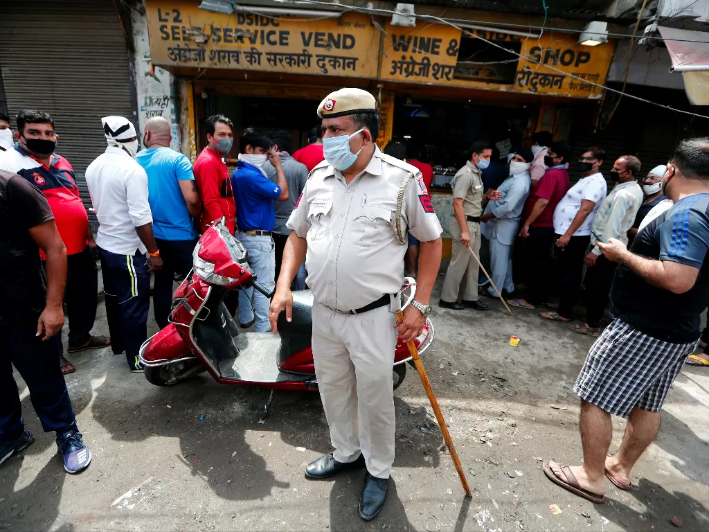 Seorang petugas polisi yang mengenakan topeng pelindung berdiri di luar sebuah toko ketika orang-orang mengantri untuk membeli minuman keras selama penguncian nasional yang diperpanjang untuk memperlambat penyebaran penyakit COVID-19, di New Delhi. (Photo
