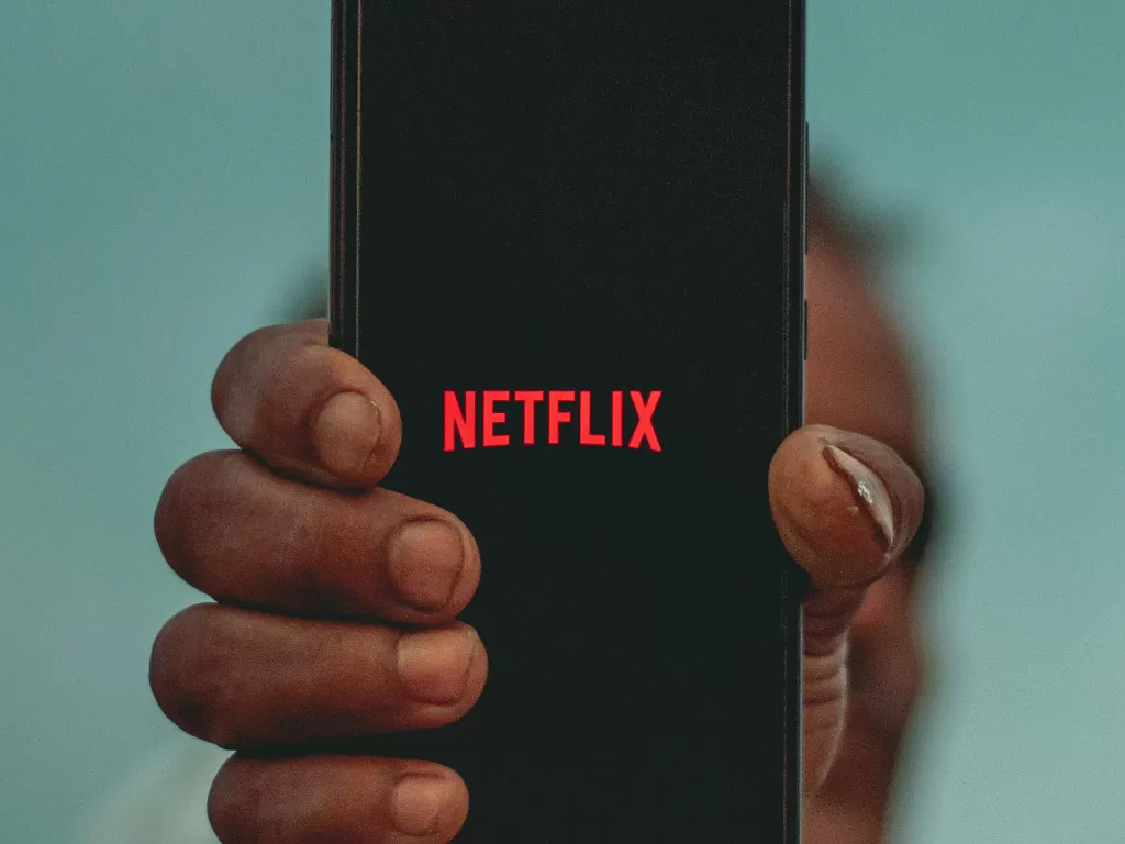 Ilustrasi seseorang sedang memegang smartphone dengan logo Netflix (photo/Unsplash/Sayan Ghosh)