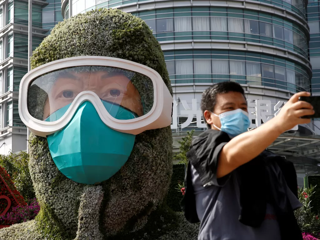 Seorang pria yang memakai masker wajah mengambil foto narsis di depan instalasi bunga yang menampilkan seorang pekerja medis dalam perang melawan penyakit virus corona (REUTERS/Tingshu Wang)