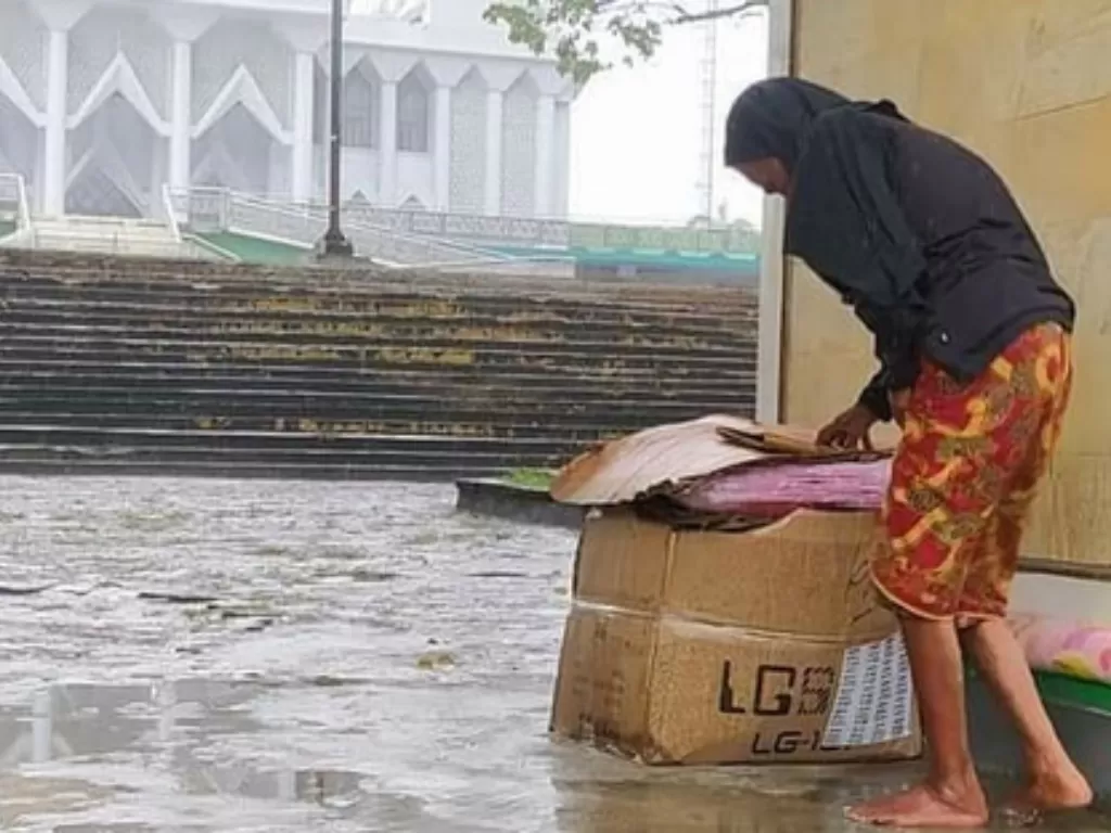 Seorang nenek yang sedang mengeringkan genangan air hujan yang mengguyur gerbang masjid yang digunakanya sebagai tempat tinggal (Foto: @viralterkini99)