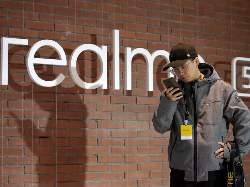 Seorang pria sedang berdiri di delam logo Realme (photo/REUTERS/Jason Lee)