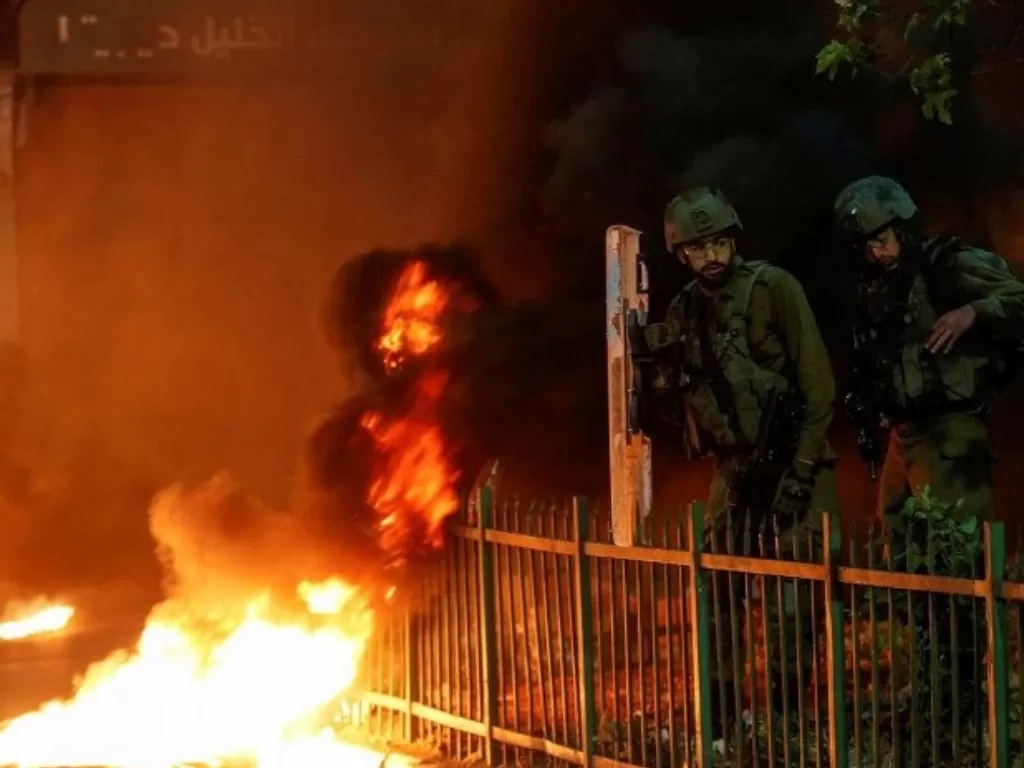 Tentara Israel berdiri di samping pembakaran ban saat warga Palestina mengambil bagian dalam protes anti-Israel atas ketegangan di Yerusalem, di Hebron di Tepi Barat yang diduduki Israel, Senin (26/4/2021). (REUTERS/Mussa Qawasma)