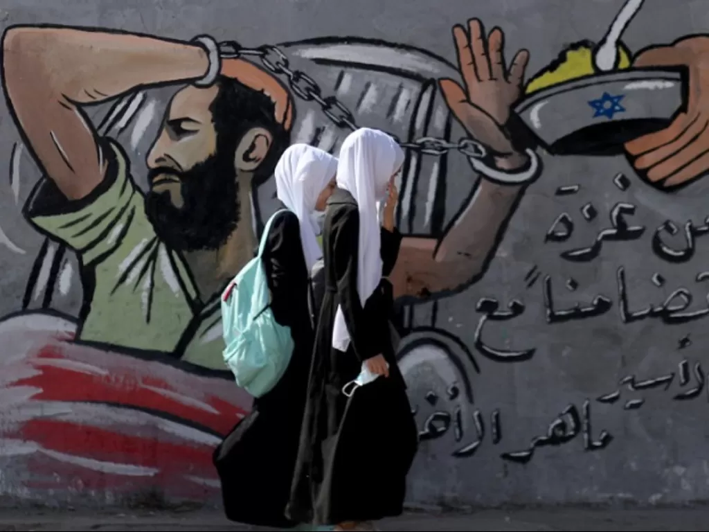 Pelajar Palestina berjalan melewati mural bergambar tahanan asal Palestina yang ditahan Israel dan menjalani mogok makan. (REUTERS/MOHAMMED SALEM)