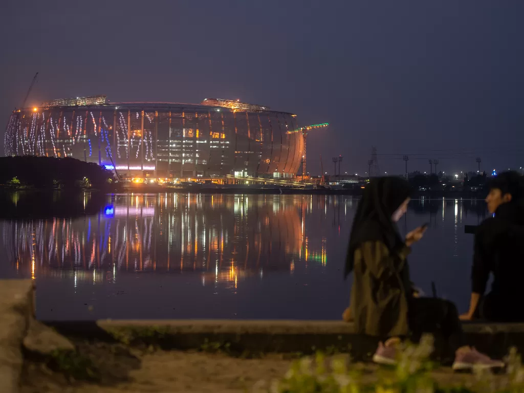 Dua warga duduk di tepi Waduk Cincin dengan latar belakang pembangunan Jakarta International Stadium (JIS) di Papanggo, Tanjung Priok, Jakarta Utara, Rabu (9/3/2022). (ANTARA FOTO/Aprillio Akbar)