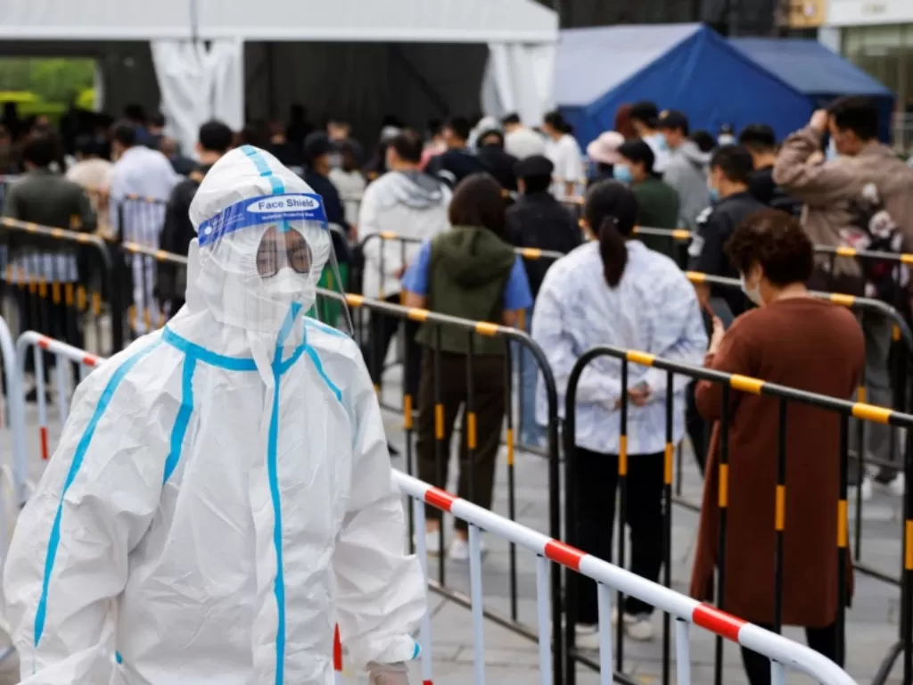 Seorang pekerja yang menggunakan APD berjalan di samping orang-orang yang mengantre untuk mengambil tes asam nukleat di tempat pengujian darurat COVID-19 di Beijing, China, 25 April 2022. (REUTERS/Carlos Garcia Rawlins)