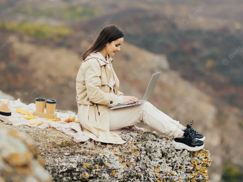 Seorang perempuan bekerja dengan laptopnya dari atas puncak gunung. (Freepik)