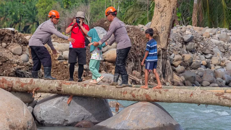 Personel kepolisian membantu seorang warga perempuan menyeberangi sungai melalui batang pohon di tengah bebatuan dan material sisa bencana hidrometeorologi. (Dok. Istimewa)