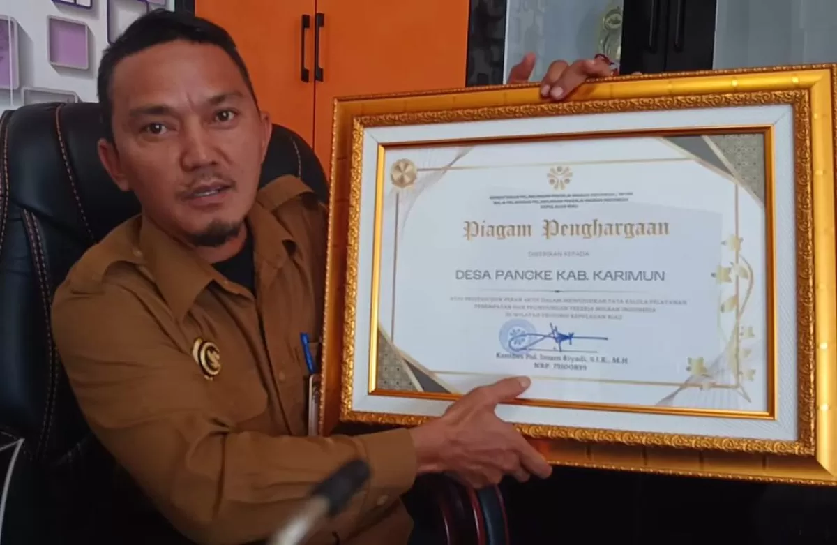 Junaidi Kepala Desa Pangke, Kecamatan Meral Barat, Kabupaten Tanjung Balai Karimun saat berada di ruangan kerja sembari melihatkan piagam prestasi yang di raih.