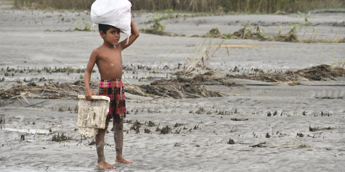 Seorang anak laki-laki membawa karung dan wadah melintasi lahan berlumpur di Kurigram, Bangladesh. Kondisi tersebut menggambarkan kerentanan masyarakat terhadap perubahan cuaca dan tekanan lingkungan yang memengaruhi kehidupan sehari-hari di kawasan tersebut. (Pexels/Masudar Rahman)