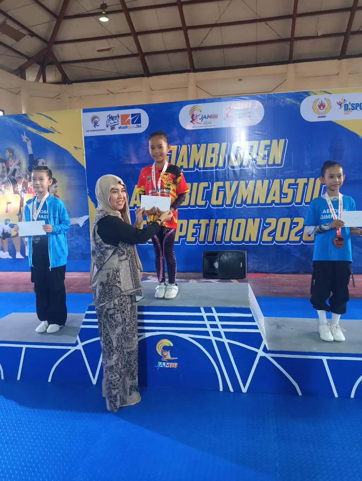 Salah satu atlet Persani Kota Payakumbuh berseragam merah-hitam berdiri di podium juara pertama menerima penghargaan dalam Jambi Open Aerobic Gymnastics Competition 2026 di Gedung Persani Provinsi Jambi, 24–26 April 2026, disaksikan atlet peraih perak dan perunggu dari kontingen lain.