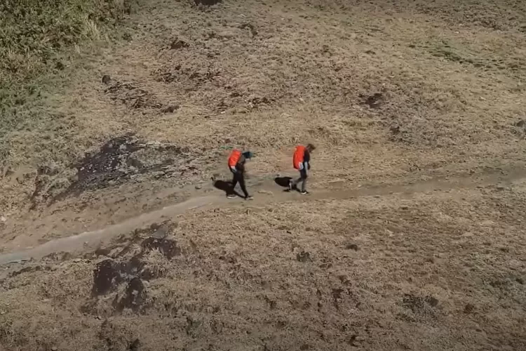 Gambar ini memperlihatkan dua pendaki berjalan di jalur setapak pegunungan dengan ransel berpenutup oranye, melintasi area berumput kering dalam suasana cerah. (Foto/YouTube - @BodinkArtventure.)